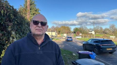 A bald man wearing tinted glasses and a blue 3/4 zip jumper stands in front of a busy crossroads. Behind him cars are driving on the road whiilst other wait at the junction to drive on.