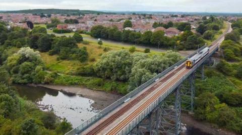 A two carriage train crosses a railway viaduct with a river below. IT's an aerial shot with a town behind