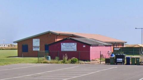 Redcar Athletic's grounds on Green Lane. A red building with a slanted roof stands in front of a larger clubhouse. Football pitches surround the buildings on three sides, while a car park has been built on the other.
