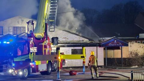 A fire engine at the scene of a white painted brick building. A fire fighter stands next to the vehicle with their back towards the camera. There is a plume of smoke coming from the building.