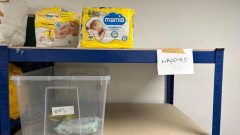 Two shelves, one with two packs of nappies on, and the other with two packs of baby wipes on. There is visible empty space on both shelves. 