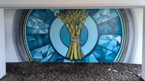 A large mural painted under a bridge, It shows a large blue coloured circle with a bundle of asparagus in the middle. The ground at the foot of the mural is strewn with rocks and stones.