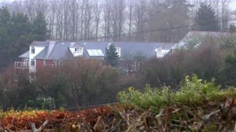 Holmesley Care Home, pictured from a distance. The home is a multi-storey building with a red brick exterior surrounded by trees.