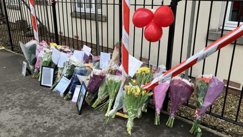 Dozens of bunches of flowers placed against a set of railings on London Road in Chippenham. The flowers include tulips, roses and carnations and some have messages pinned to them. Above the bunches of flowers are three red balloons tied to the railings. 