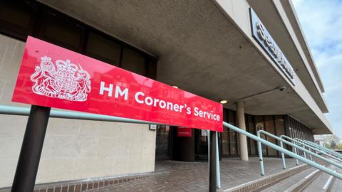 A red sign at the entrance to Essex Coroner's Court. The sign reads "HM Coroner's Service". Brick steps lead to a revolving door.
