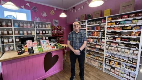 The image shows the interior of a cheerful sweet shop with pink-painted walls and a playful, festive atmosphere. Shelves are stocked with a variety of confectionery, and a wooden-topped counter featuring a large heart design on the front displays boxes of sweets, jars, and other treats. Behind the counter, glass jars are filled with assorted sweets, and a Jelly Belly jelly bean display is visible on the right-hand side. Decorative touches such as bunting and circular signs reading "LOVE YOU," "HAPPY BIRTHDAY," and "BE MINE" hang from the ceiling, adding to the celebratory feel. A person wearing a checked shirt and black trousers stands in front of the counter with their hands clasped together.