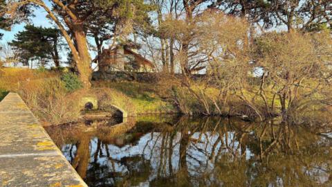 Photo of the reservoir in Guernsey. The sun is shining down on the water and there are blue skies.