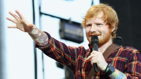 A ginger haired singer wearing a red checked shirt singing into a mic with tattoos up his arm.
