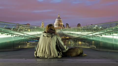 A homeless man sitting on the ground with his back to the camera, wearing a grey hoodie with a dog lying by his side. He is facing the dome of St Paul's Cathedral and on both sides of him are illuminated glass walkways