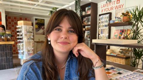 Woman with brown hair wearing denim shirt and star earrings sits in shop with racks of bracelets behind her and trays of beads