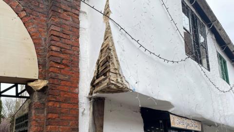 The exterior of a damaged building. A section of red brickwork, arranged in a traditional staggered pattern, is clearly visible. There is a stone block embedded partly in the brick column, protruding slightly. Next to it appears to be a wooden panel or board placed over an opening or archway.