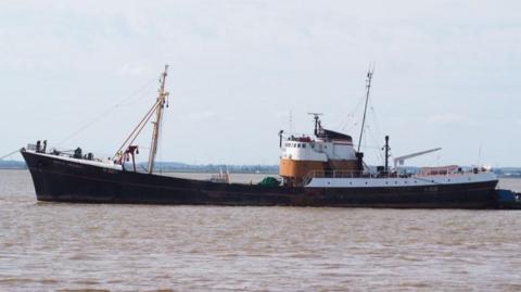 A large ship in water. It is black with a white and brown bridge and two masts.
