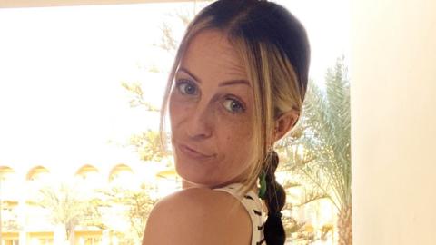 Kirsty Buchan looks over her shoulder. She has blonde and brown hair tied in a plait and is wearing a white and black striped vest top.