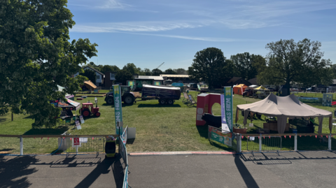 A field includes stalls and a tractor. There are two green banners either side of the entrance to the field near the bottom of the image. There are trees on the left and in the background.