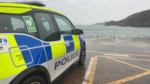 The side of a Jersey police van. It is parked beside the sea on a cloudy day.