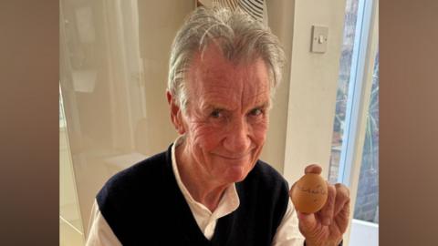 Sir Michael Palin sits at a kitchen table and holds an egg in one hand that he has signed with a pen. He has short grey hair and wears a white shirt with a black vest over the top.