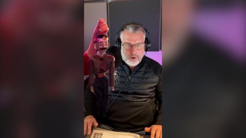 A wooden puppet meant to look like Hamlet has been edited next to Andy Collins. He has glasses on and a beard, his hair is grey. He looks shocked. 