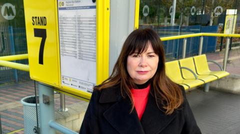 Anneliese Midgley, who has long brown hair with a fringe and wears a black trench coat over a red jumper, stands by a glass bus shelter with a sign reading 'Stand 7'.