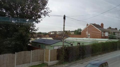 A group of green caravans behind a fence in a residential area with houses behind and a road in front.