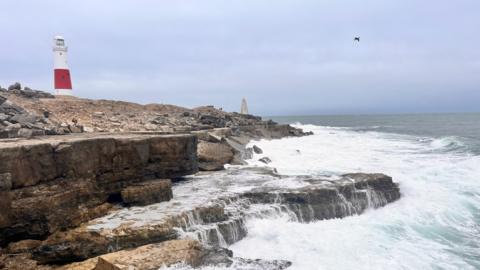 Waves crash over rocks on the Dorset coast. The sky over head is grey. On the headland you can see the red and white Portland Bill lighthouse