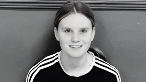 Black-and-white picture of Holly. She is sitting with her back against a wall and is smiling at the camera. Her long hair is in a ponytail and she is wearing a black top with white stripes down the shoulders.