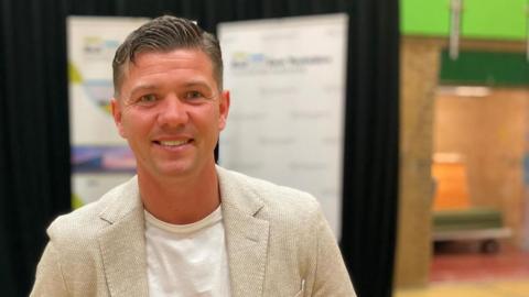 Luke Campbell smiles into the camera after winning the mayoral election in Hull and East Yorkshire. He is wearing a beige blazer with a white t-shirt. He is standing in front of a stage that includes a podium and a black background.