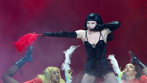 Lady Gaga wears a black one-piece and a black wig as she performs on stage holding a feather boa 