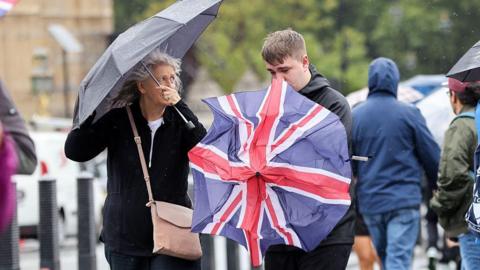 A woman under a plain umbrella and a man struggling to put up a Union Flag umbrella in windy conditions