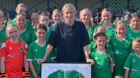 Leah Williamson holding a framed shirt while posing with a team of young girls