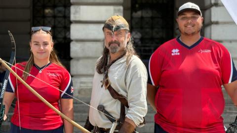 A man and woman in red Team GB archery sports tops stand either side of a man dressed as Robin Hood. He is wearing a cream/white coloured top and is holding a bow and arrow