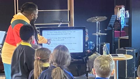 A group of children standing looking at a TV screen, a man is pointing at the screen