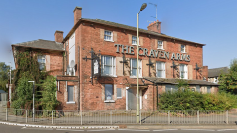 A red brick building with the words The Craven Arms at the top. Trees surround the building, alongside some street fencing