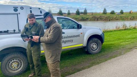 Two men, wearing dark green clothes, the man on the right is wearing a black cap. The man on the left is holding a tablet. Behind the two of them is a van which says "Environment Agency" on it. The van is parked next to a riverbank. 