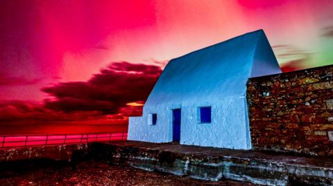 white cottage with a blue door. the sky is red and green due to the Northern Lights 