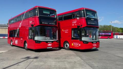 Two red double-decker buses are parked side by side in an open area. Both display the text '100% electric • zero emissions' on the front. The left bus has 'city' written on it, while the right bus includes branding for a project with Cornwall Council and partners. Additional red buses are visible in the background.

