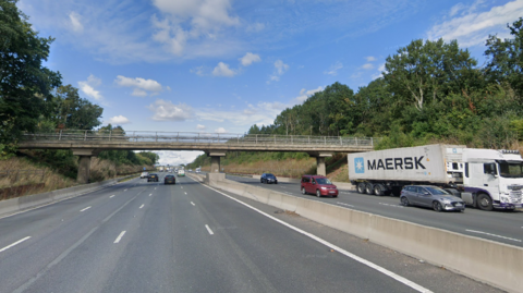 A Google street maps view of the M1dual-carriageway heading south between Northampton and Milton Keynes. There are a number of cars on either side of the road, and trees on either side of the road too. 