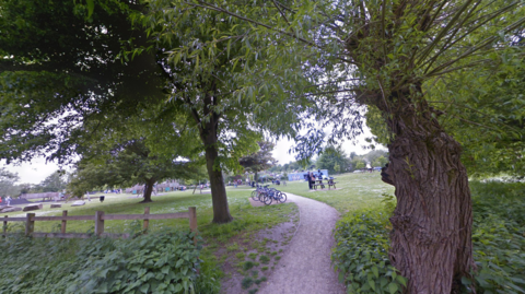 The entrance to Toddler's Cove playground in Whitehall Road, Canterbury, showing a path through trees, with bikes standing in the foreground and play equipment in the background.