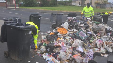 Two men in high vis jackets working on a large pile of rubbish, which has been emptied on to a road. Black wheelie bins surround the pile. In the background on the other side of the road is a bus stop and a pedestrian in the distance.