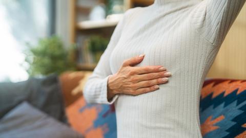 Close-up of person with their arm raised and a flat palm on the lower side of their breast. There is a sofa with a geometric pattern on it in the background.