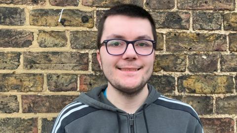 Callum has short dark brown hair, black-frame glasses and is wearing a grey Adidas-branded hooded top. There is a brick wall behind him.