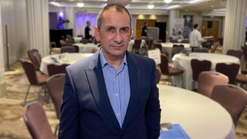 Murray Norton wears a blue striped shirt and blazer. He is standing in a room with round tables and beige chairs.