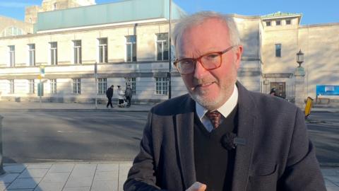 A white man with white hair, beard and glasses is in a suit outside a white building in Southampton looking down the end of the camera.
