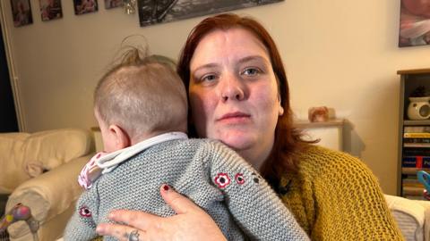 A woman in a yellow knitted jumper holding a baby in a grey knitted jumper against her shoulder.