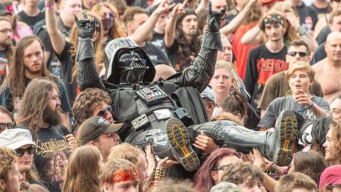 A person dressed in a black Darth Vader costume with helmet and gloves crowd surfs over people at a heavy metal music festival.