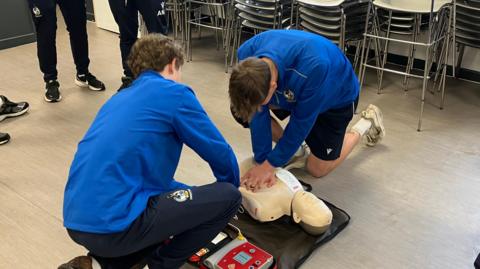 Two boys knelt down next to a medical training dummy. One has his hands locked on the dummy's chest - performing CPR. They are wearing blue jumpers and trousers with Bristol Rovers logos on them. 