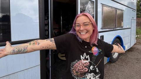 Lyndzi Hargrave smiling with her arms outstretched in front of a double decker bus with a light blue exterior.