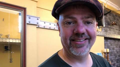 Des Reynolds is pictured with a goatee beard and a flat cap is smiling at the camera. He is wearing a black T-shirt and is standing indoors in a cafe with cream-coloured walls.