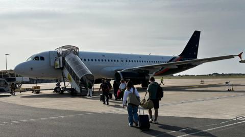 A Titan aircraft, with passengers going up a glazed stairway. It's a stubby jet aircraft with a white body and a blue wing and tail. 