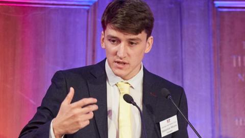 Sebastian Moore is clean shaven, has short brown hair and is wearing a dark blue suit with a white shirt and yellow tie.