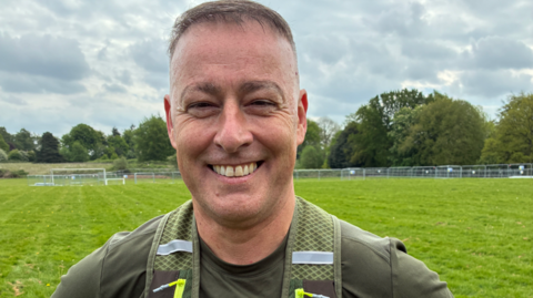 A man with short brown hair and a green top in front of a large grassy area